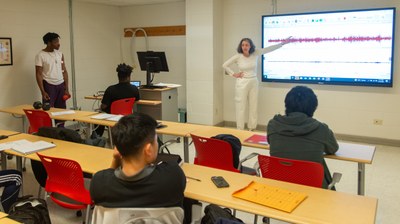 Dr. Gali Werber-Zion, a professor, infront of a classroom with students, pointing at a screen that resembles soundwaves, which are the result of electromyography